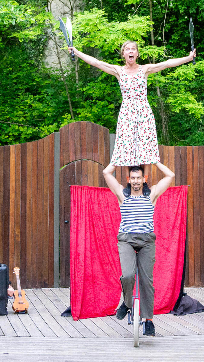 Artist from Cirque Les Dudes perched on a colleague's shoulders, juggling knives during the 'Stories in the City!' show on an outdoor wooden stage.