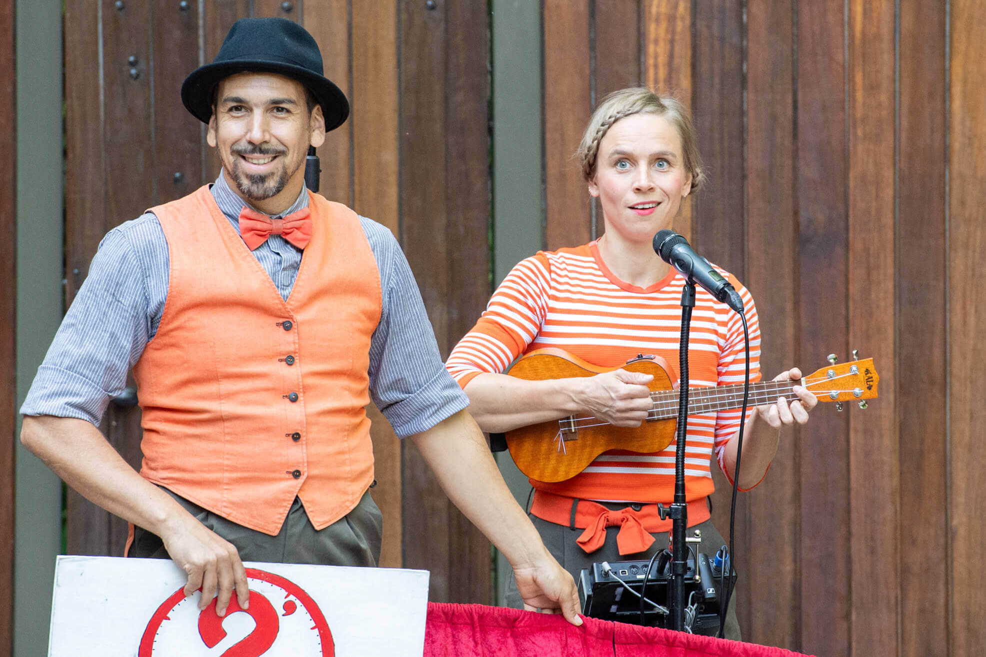 Man with hat, orange vest and bow tie, woman in orange stripes playing the ukulele, street performance, wooden background.