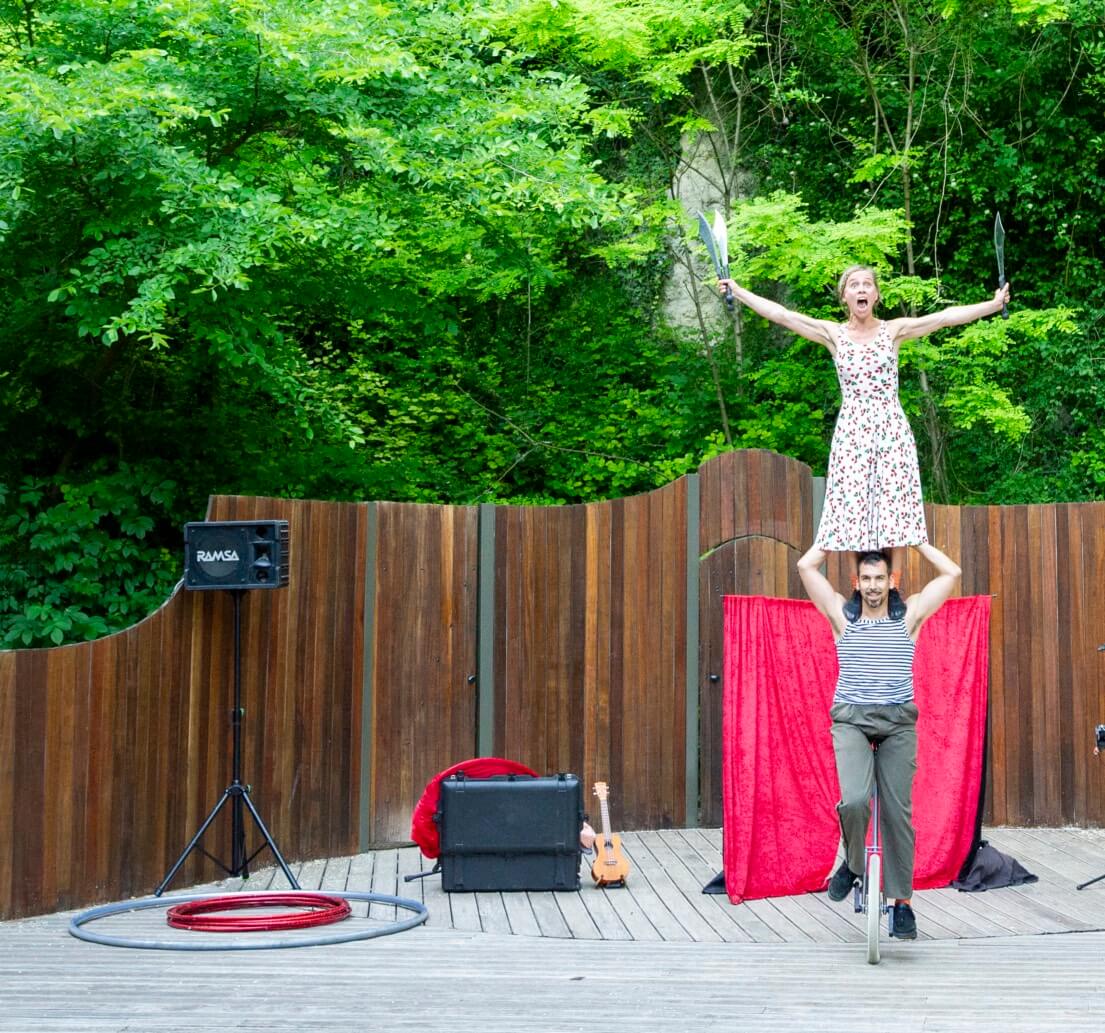Artiste du Cirque Les Dudes juchée sur les épaules d'un collègue, jonglant avec des couteaux durant le spectacle Stories in the City! sur une scène extérieure en bois.